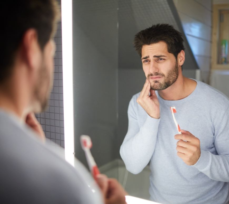 Man holding toothbrush in one hand and holding his cheek in pain in the other